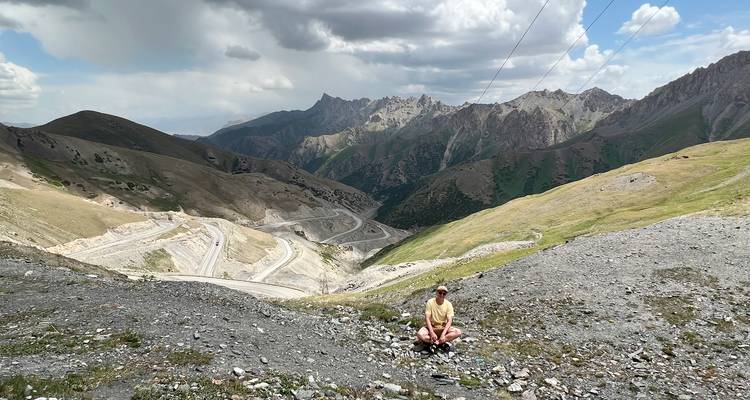 Alleinreisender ruht an einem Bergpass mit Blick auf gewundene Alpenstraßen und zerklüftete Gipfel.