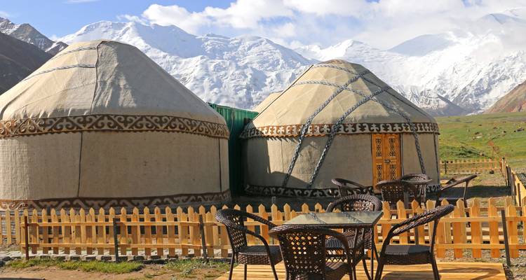 Traditionelle Filzjurten stehen vor schneebedeckten Gipfeln mit Holzzaun und Sitzgelegenheiten.