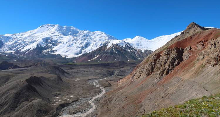 Raue alpine Tal mit einem Gletscherfluss unter schneebedeckten Gipfeln und blauem Himmel.