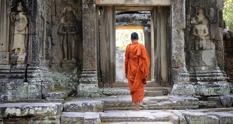 Monje budista con túnicas naranjas caminando a través de la entrada de un templo de piedra antigua adornada con deidades talladas.