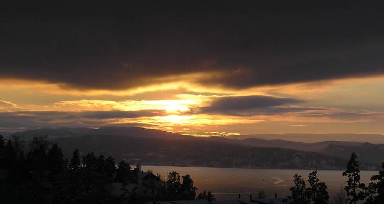 Coucher de soleil doré perçant à travers les nuages sombres au-dessus de l'Oslofjord et des collines lointaines.