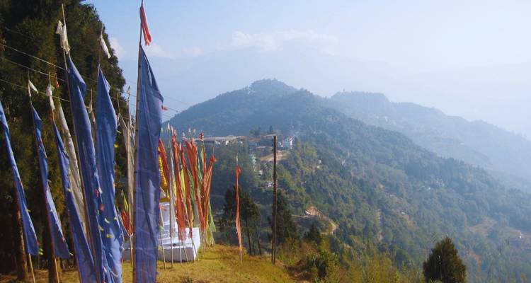 Bunte Gebetsfahnen flattern neben einem Grat mit nebligen Himalaya-Vorbergen dahinter.
