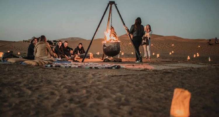 Les voyageurs sont assis autour d'un feu de chaudron suspendu sous le crépuscule du désert avec des lanternes parsemant le sable.