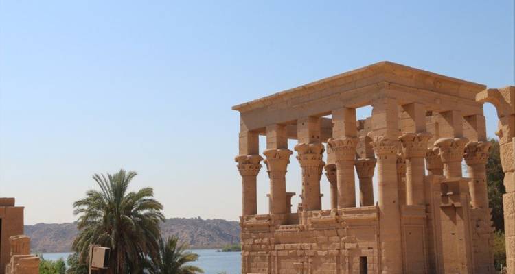 Ancient sandstone columns of a riverside temple with palm trees and the Nile in the background.