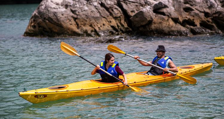 Two travellers paddling a bright yellow kayak in clear turquoise bay water below rocky cliffs
