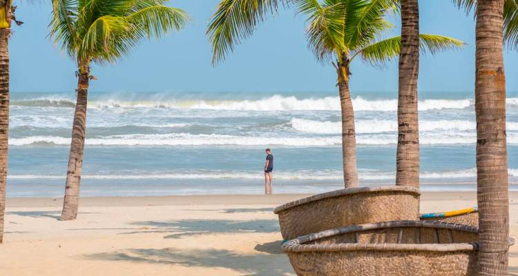 Quiet tropical beach framed by palm trees and round basket boats with a lone walker near surf