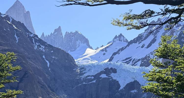 Vista dramática de las puntiagudas agujas del Fitz Roy y un glaciar colgante enmarcados por árboles verdes.