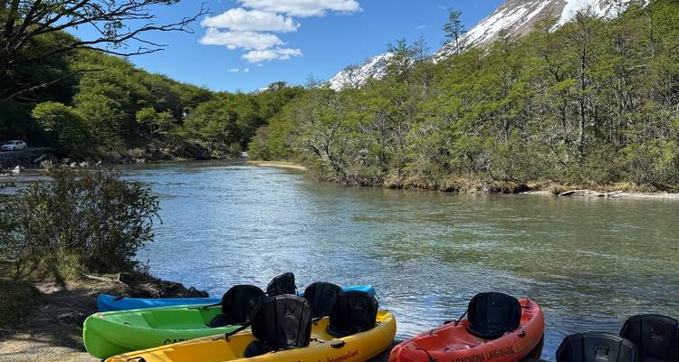 Kayaks coloridos descansan en la orilla tranquila de un río bordeada por bosque y picos nevados en la distancia.
