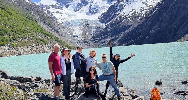 Pequeño grupo posa en la orilla rocosa del lago con lago glaciar turquesa y picos nevados detrás.