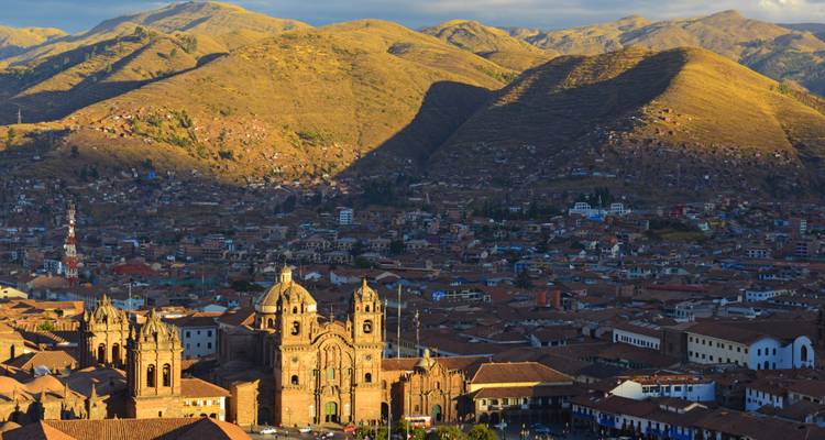 Golden late-afternoon sun over Cusco rooftops with cathedral and surrounding Andes.