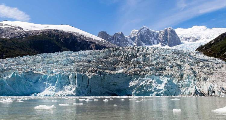 A vast blue glacier calves into an icy fjord backed by snow-capped peaks under a bright sky.
