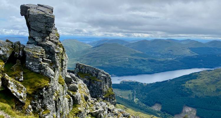 Afloramientos rocosos dentados dominan amplios valles de las tierras altas y un lago reluciente bajo nubes dispersas.
