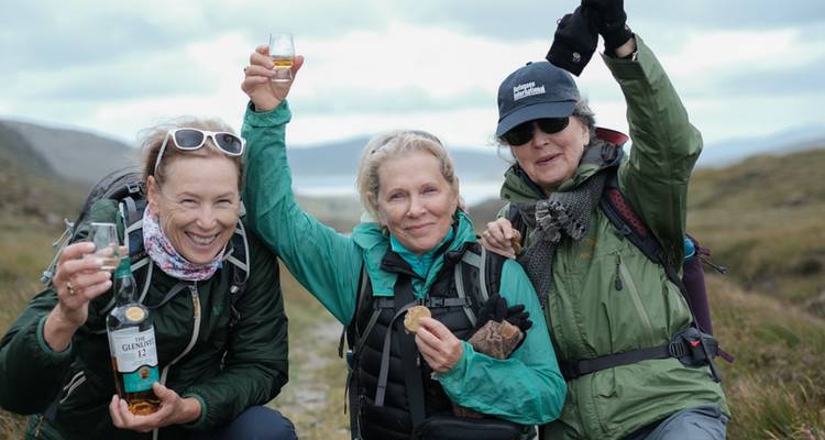 Tres excursionistas levantan bebidas en celebración en un sendero de montaña azotado por el viento, vistiendo equipo de montañismo y mochilas.