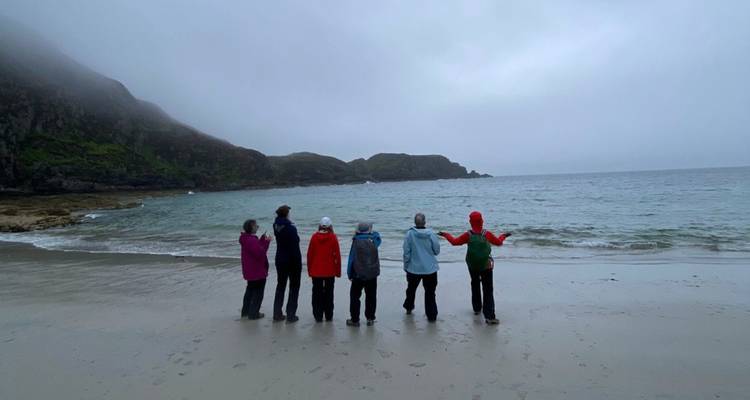 Seis personas están de pie mirando hacia las olas en una playa brumosa y desierta con promontorios rocosos en la distancia.