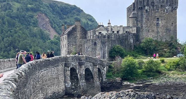 Los turistas cruzan el puente de piedra hacia un castillo histórico situado contra acantilados verdes de las tierras altas.