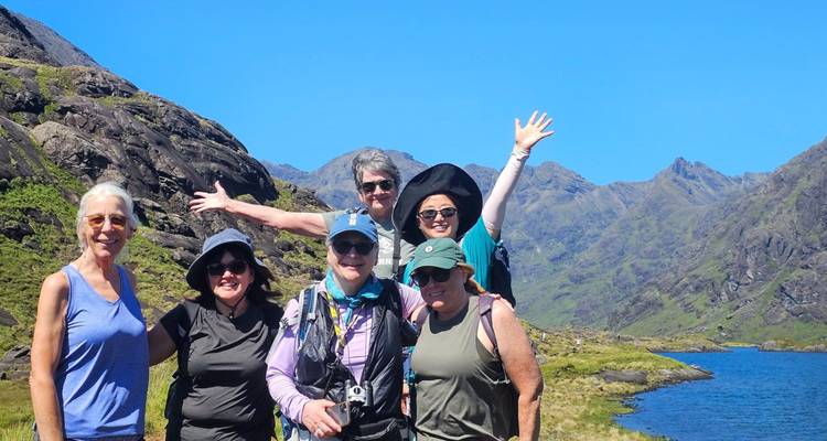 Un grupo de senderismo alegre posa junto a un lago de montaña con picos escarpados elevándose detrás bajo un cielo azul despejado.