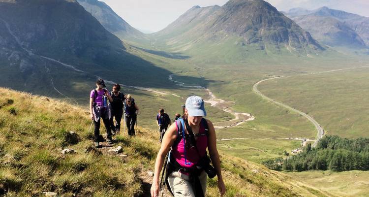 Una fila de excursionistas asciende por una empinada ladera cubierta de hierba en un dramático valle montañoso en forma de U con un río serpenteante muy abajo.