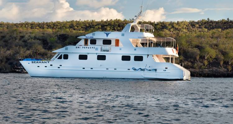 White live-aboard yacht Seaman Journey anchored off rocky Galápagos island shore.