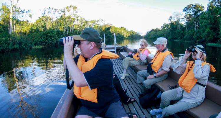 Kleines Motorboot von Reisenden mit Schwimmwesten beobachtet Vögel von einem Holzdeck auf ruhigem Fluss.