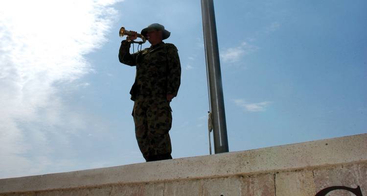 Corneta silueteada en uniforme militar toca una trompeta junto a un asta de bandera contra el cielo.