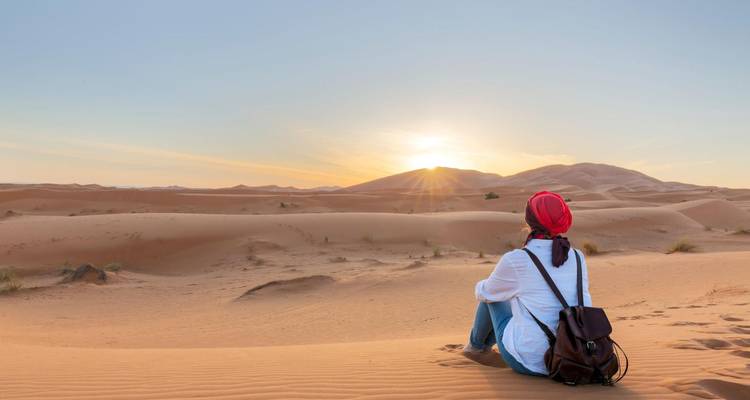 Un viajero con pañuelo en la cabeza se sienta sobre dunas doradas observando el sol ponerse sobre el Sahara.