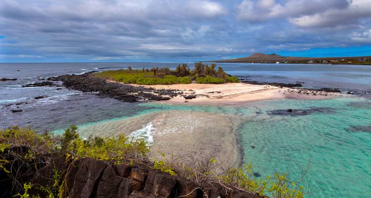 Kleine sandige Insel, umgeben von türkisfarbenem Wasser und schwarzem Vulkangestein auf den Galápagos unter einem teilweise bewölkten Himmel.