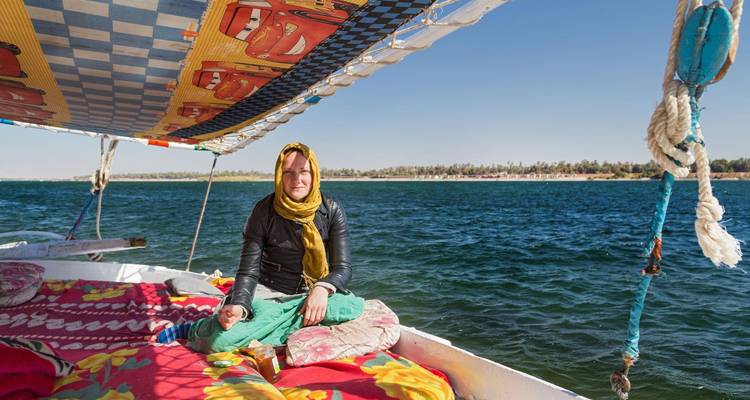 Reisender sitzt entspannt auf bunten Kissen an Bord einer Feluke, die auf den blauen Gewässern des Nils segelt.