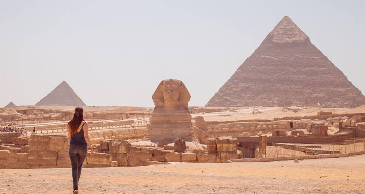 Ein Besucher geht auf die Große Sphinx zu, während sich die Pyramiden von Gizeh in der dunsigen Wüstenkulisse erheben.