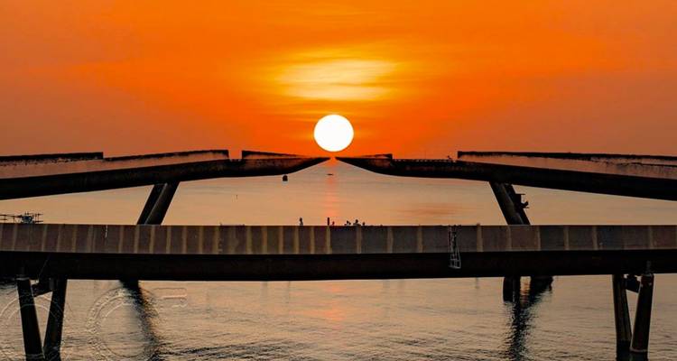 Levendige oranje zonsondergang omlijst door twee gebroken brugspannen boven kalm zeewater