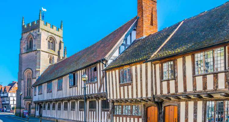 Des bâtiments Tudor à colombages et un clocher d'église bordent une rue ensoleillée de Stratford-upon-Avon.