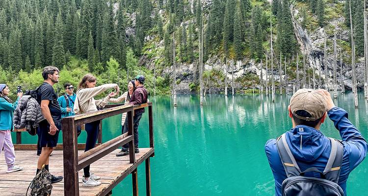 Tourists on a wooden platform overlooking the turquoise, tree-filled Kaindy Lake surrounded by conifer forest.