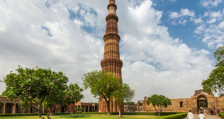 La alta torre Qutub Minar de arenisca roja se alza contra un cielo parcialmente nublado con jardines alrededor.