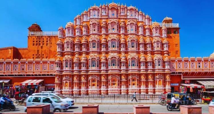 Fachada rosada intrincada del Hawa Mahal brillantemente iluminada bajo un cielo azul profundo con tráfico al frente.