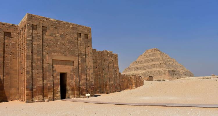 Antigua Pirámide Escalonada de Saqqara elevándose desde el desierto arenoso con edificio de entrada de piedra caliza adyacente bajo un cielo azul despejado.