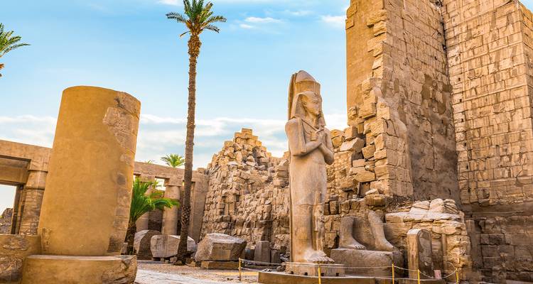 Estatua de faraón entre palmeras y ruinas en el Templo de Luxor bajo el cielo azul.