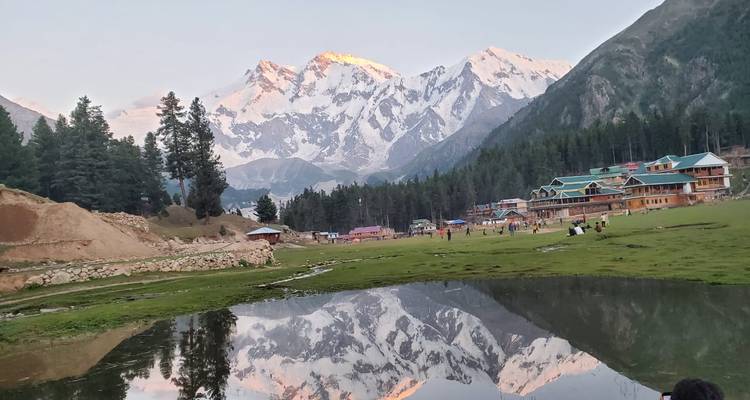 L'alpenglow illumine un sommet enneigé se reflétant dans un étang de prairie près de chalets en bois et de visiteurs dispersés.