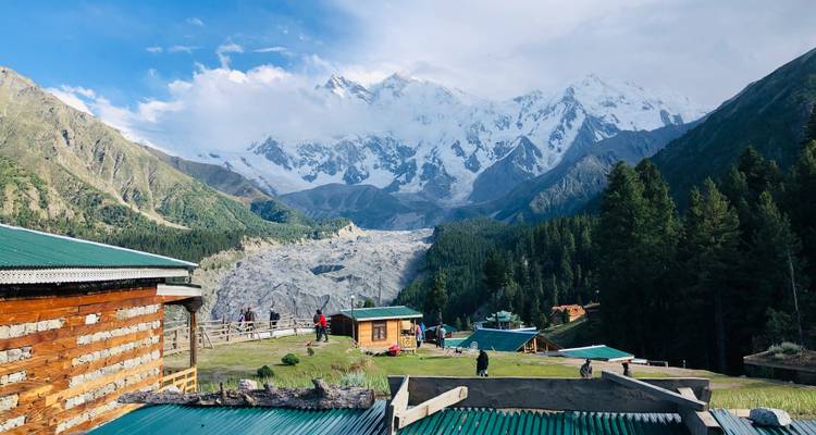 Les visiteurs se promènent devant des chalets rustiques sur une crête herbeuse surplombant un glacier et un sommet enneigé imposant.