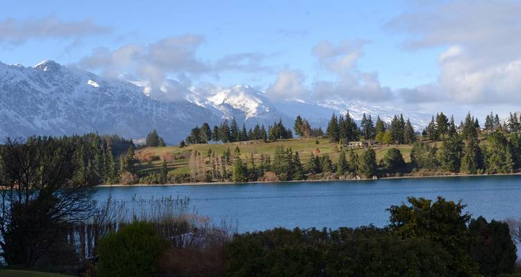 Des montagnes couvertes de neige se dressent au-dessus d'un lac bleu profond bordé d'arbres à feuilles persistantes et de pentes herbeuses.