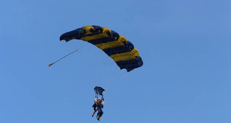 Un couple de parachutistes en tandem glisse sous un parachute bleu et jaune dans un ciel dégagé.
