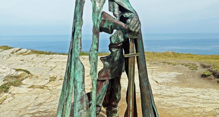 Une statue de bronze patinée d'une figure encapuchonnée tenant une épée se dresse sur une falaise côtière rocheuse.