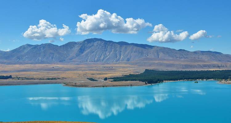 Un lac turquoise éclatant reflète des nuages cotonneux avec des collines dorées arides et des montagnes escarpées en arrière-plan.