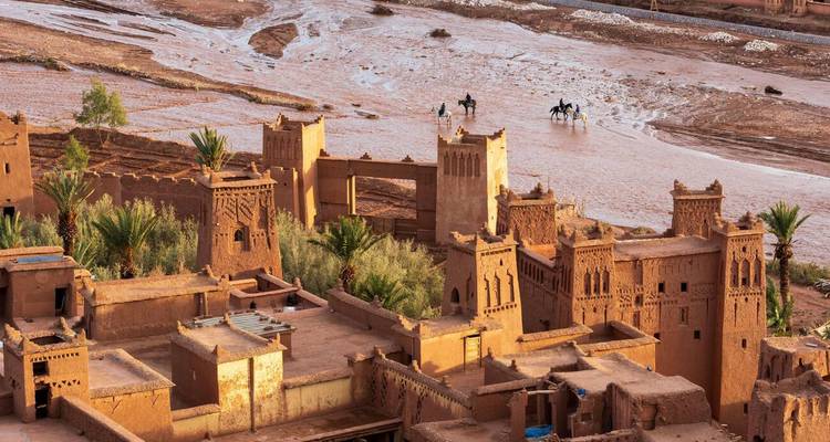 Vue aérienne du ksar fortifié en terre d'Ait Benhaddou au bord d'une rivière peu profonde avec des cavaliers qui traversent