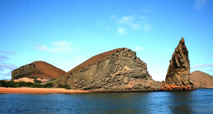 Pinnacle Rock rises sharply beside arid volcanic hills and calm blue water under a clear sky.
