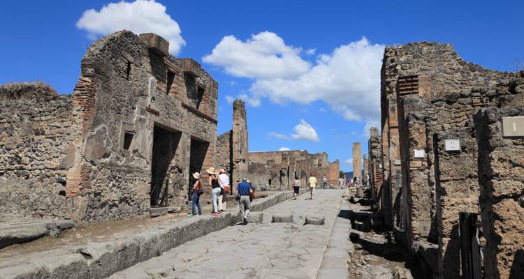 Calle de piedra con edificios en ruinas y turistas explorando la antigua Pompeya bajo cielos azules.