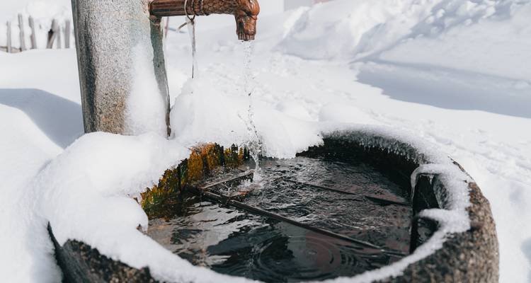 Alter steinerner Brunnen lässt Wasser in ein schneeberandetes Becken in einer stillen Winterlandschaft tröpfeln.