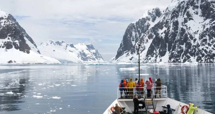 Passengers stand on a ship’s bow amid floating ice and towering Antarctic fjords.