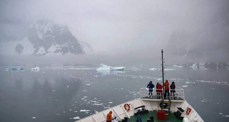 Ship navigates through foggy Antarctic waters with small icebergs; guests bundled on deck.