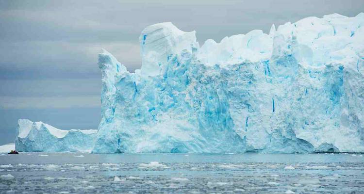 Massive blue-white iceberg towering above a calm grey sea under cloudy skies, with small bits of floating ice in the foreground.