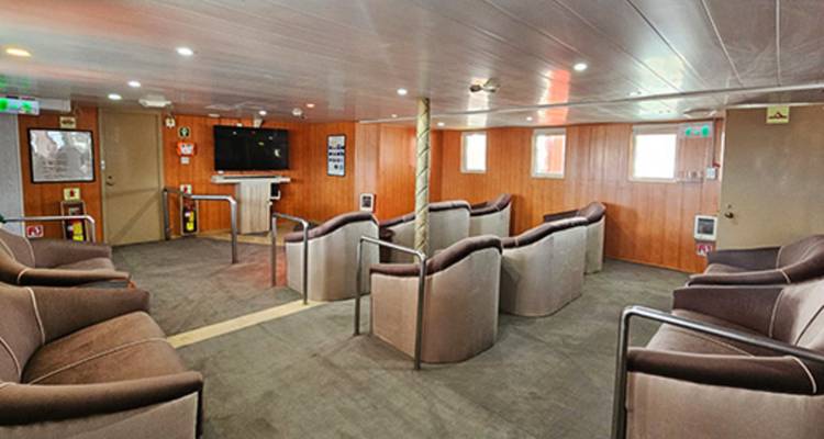 Simple shipboard lounge with cushioned armchairs facing a television and wooden paneled walls.