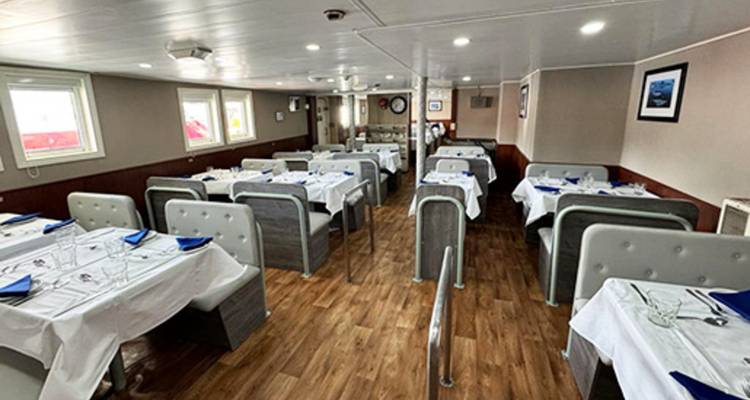 Clean onboard dining room with neatly set tables, white tablecloths and wooden flooring.